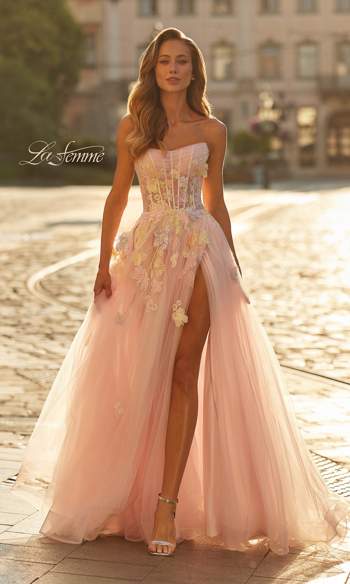 A woman stands on a sunlit cobblestone street wearing the La Femme 33432 long prom dress in light pink, featuring a strapless neckline, high slit, and floral appliqué.